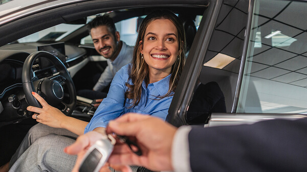 Mujer sentada en el asiento del conductor recibiendo la llave de su auto nuevo, con un hombre al fondo en el asiento del acompañante