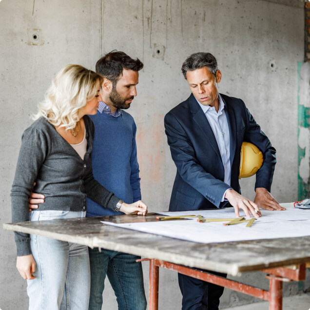 Tres personas en una obra, analizando y discutiendo un proyecto en papel sobre una mesa, mientras escuchan la explicación del ingeniero ejecutivo