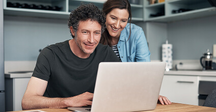 Hombre y mujer en la cocina, felices mirando la pantalla de una notebook sobre la mesa; él sentado tipeando y ella de pie apoyada en su hombro