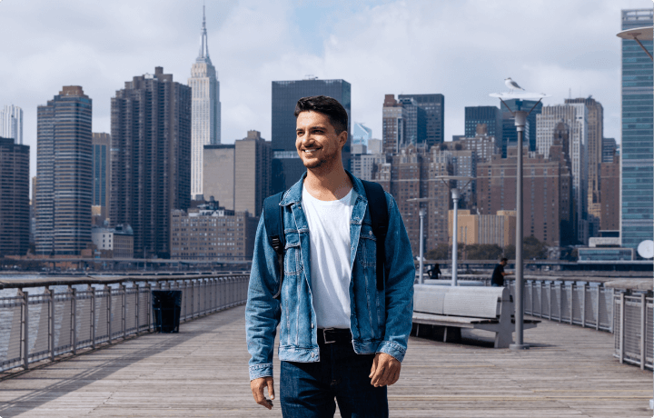 Hombre joven sonriendo y mirando al horizonte mientras camina por un puente, con la ciudad desenfocada al fondo