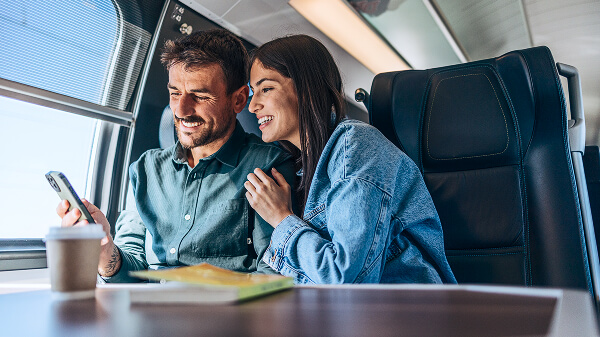 Pareja en el interior de un tren, sonriendo y mirando la pantalla del celular mientras él lo sostiene y ella se apoya en su hombro